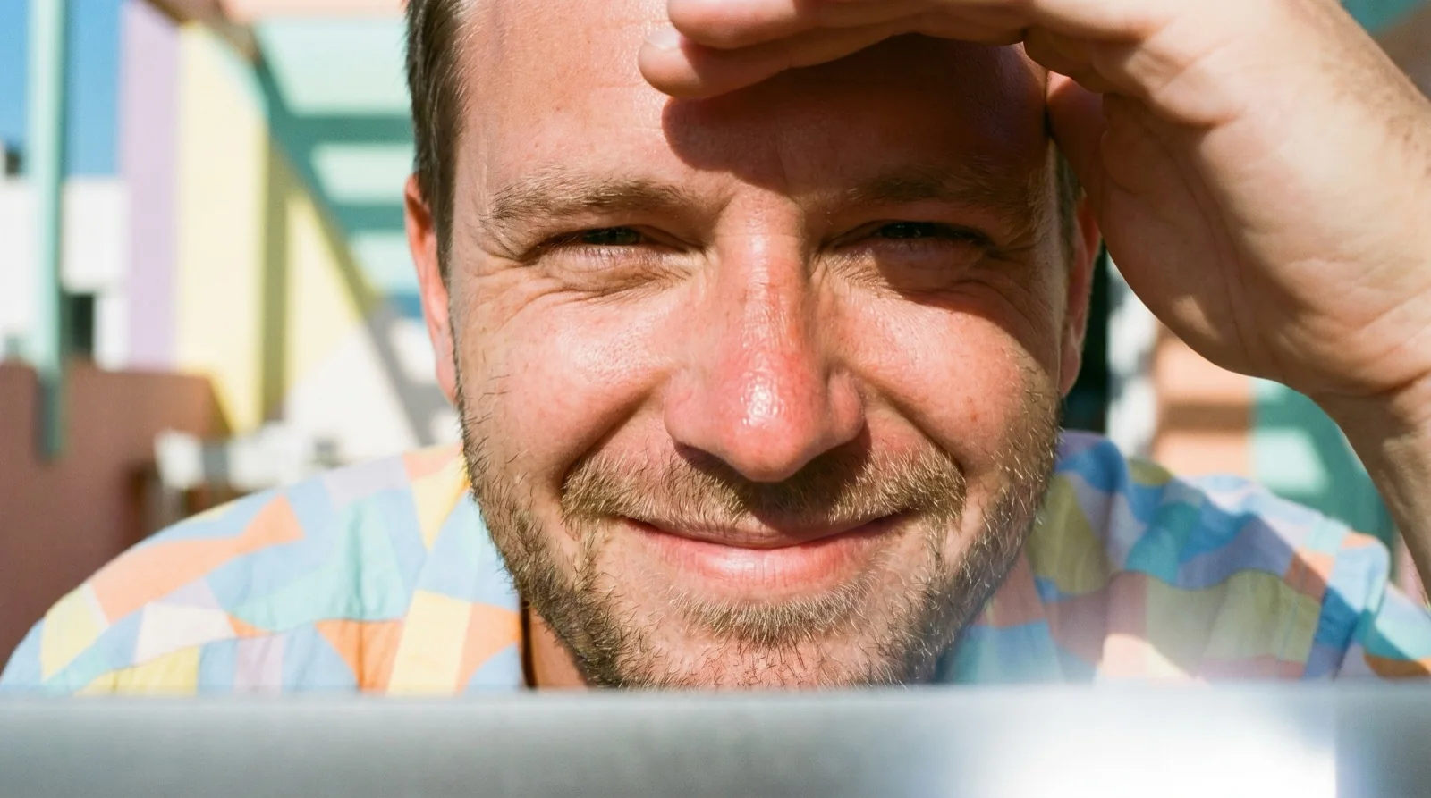 Smiling man shading his eyes with one hand, looking over the lid of a laptop in bright sun with pastel buildings behind him