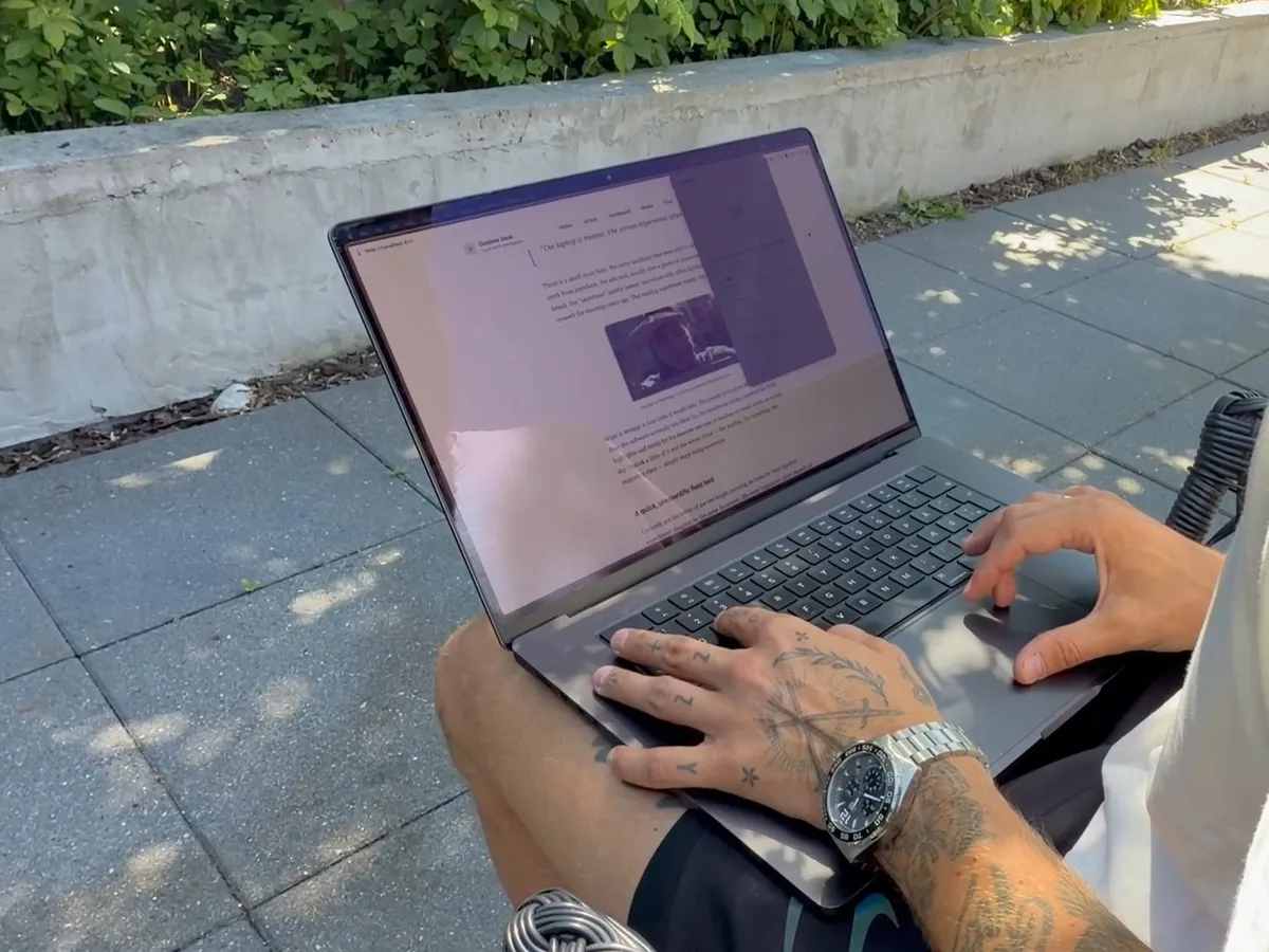 Hands typing on a 16-inch MacBook Pro on a terrace in patchy sun, with Outshine switched on and a light article page on the screen. An unedited frame from an iPhone video, July 2026