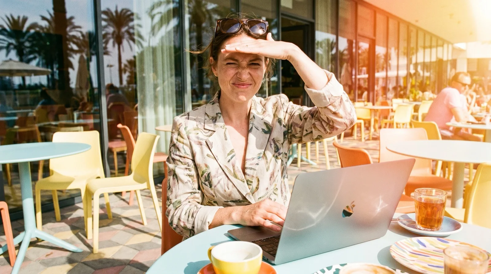 Woman at a sunny cafe terrace shading her eyes with one hand while typing on a silver MacBook